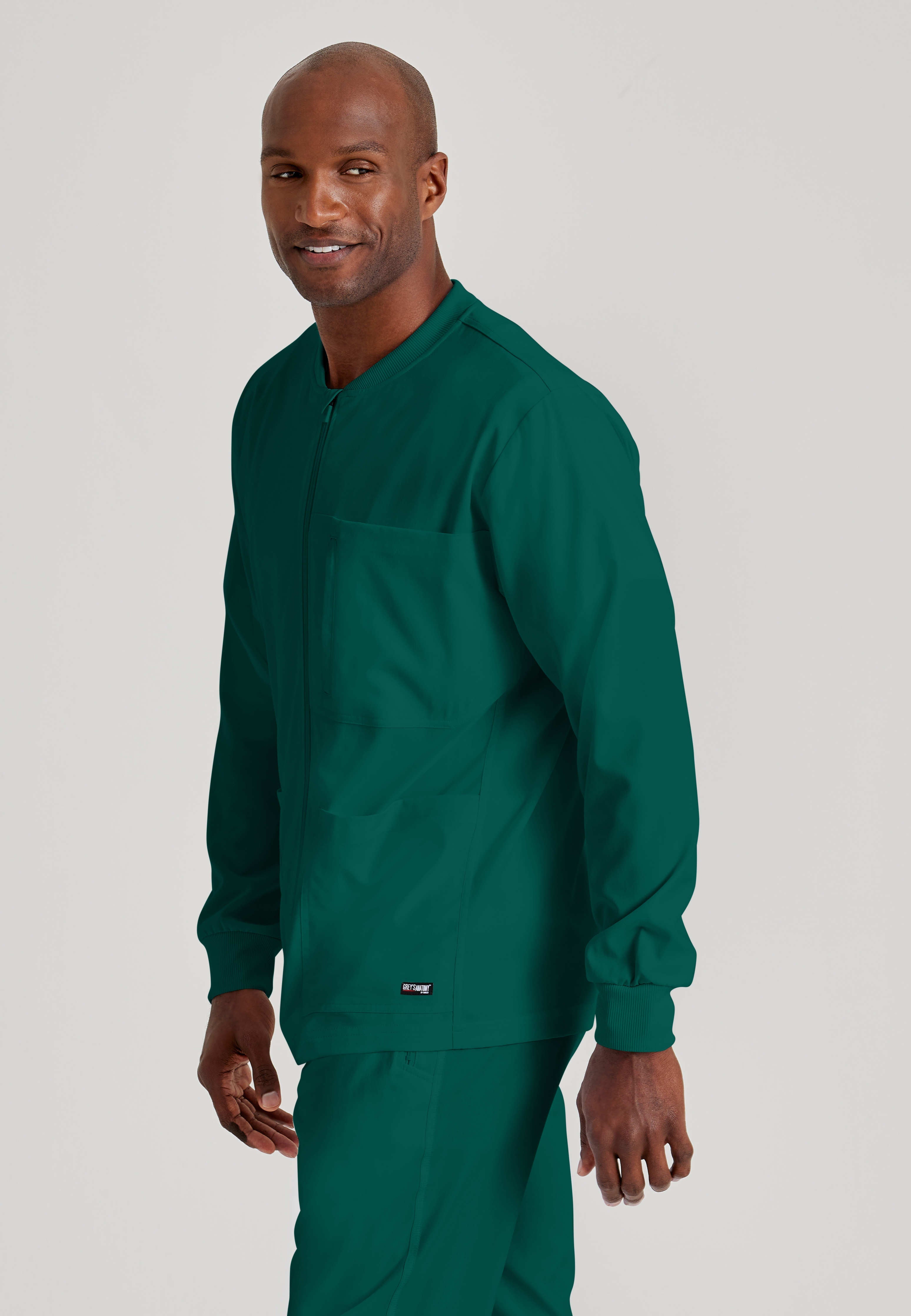 Side View of the React 5-Pocket Crewneck Warmup Scrub Jacket in Hunter Green from Grey's Anatomy™ by Barco Spandex Stretch